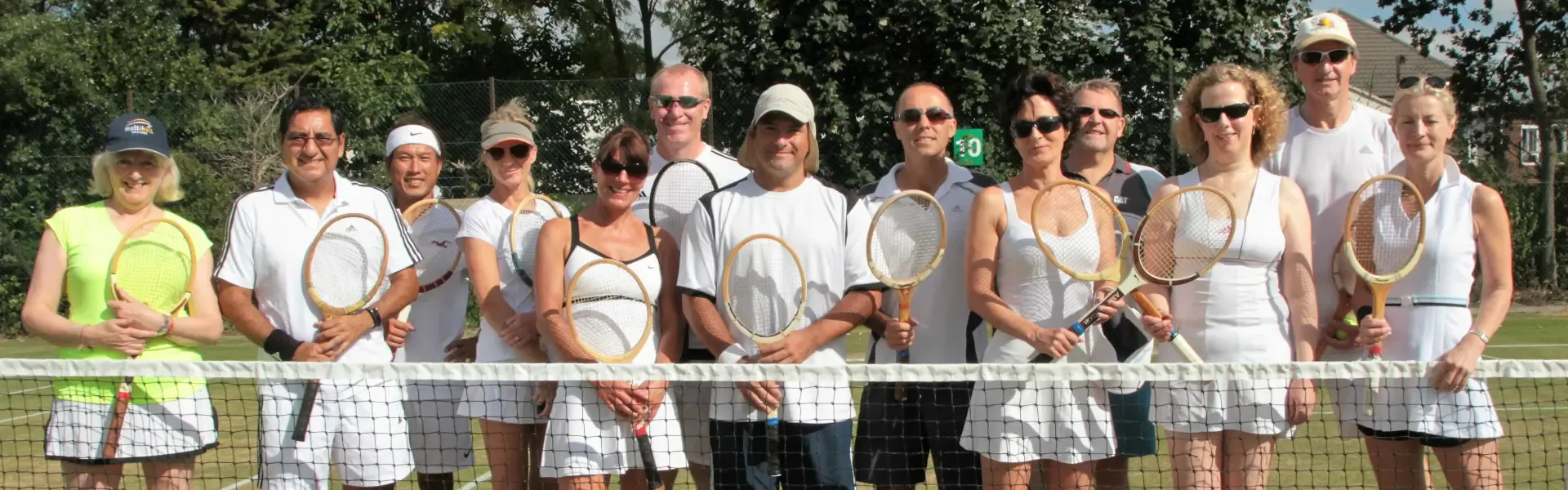 A large group of members in tennis whites holding wooden rackets, gathered at the net on the grass courts
