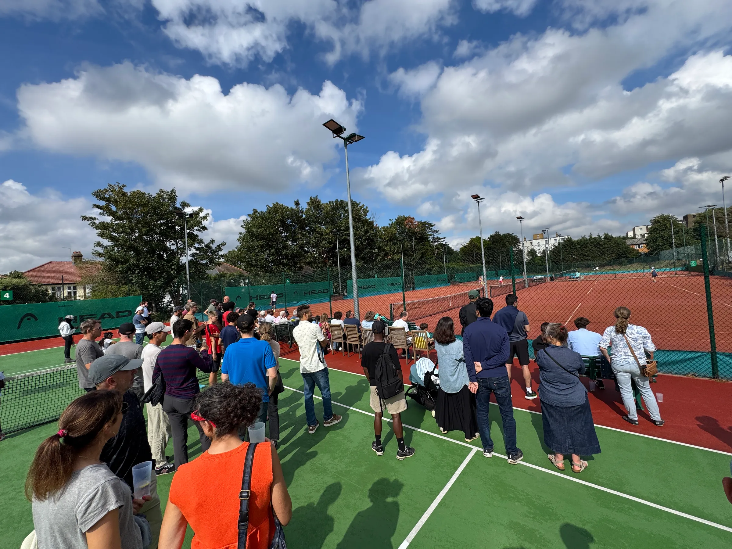 A crowd of members watching tennis beside the clay courts under a blue sky