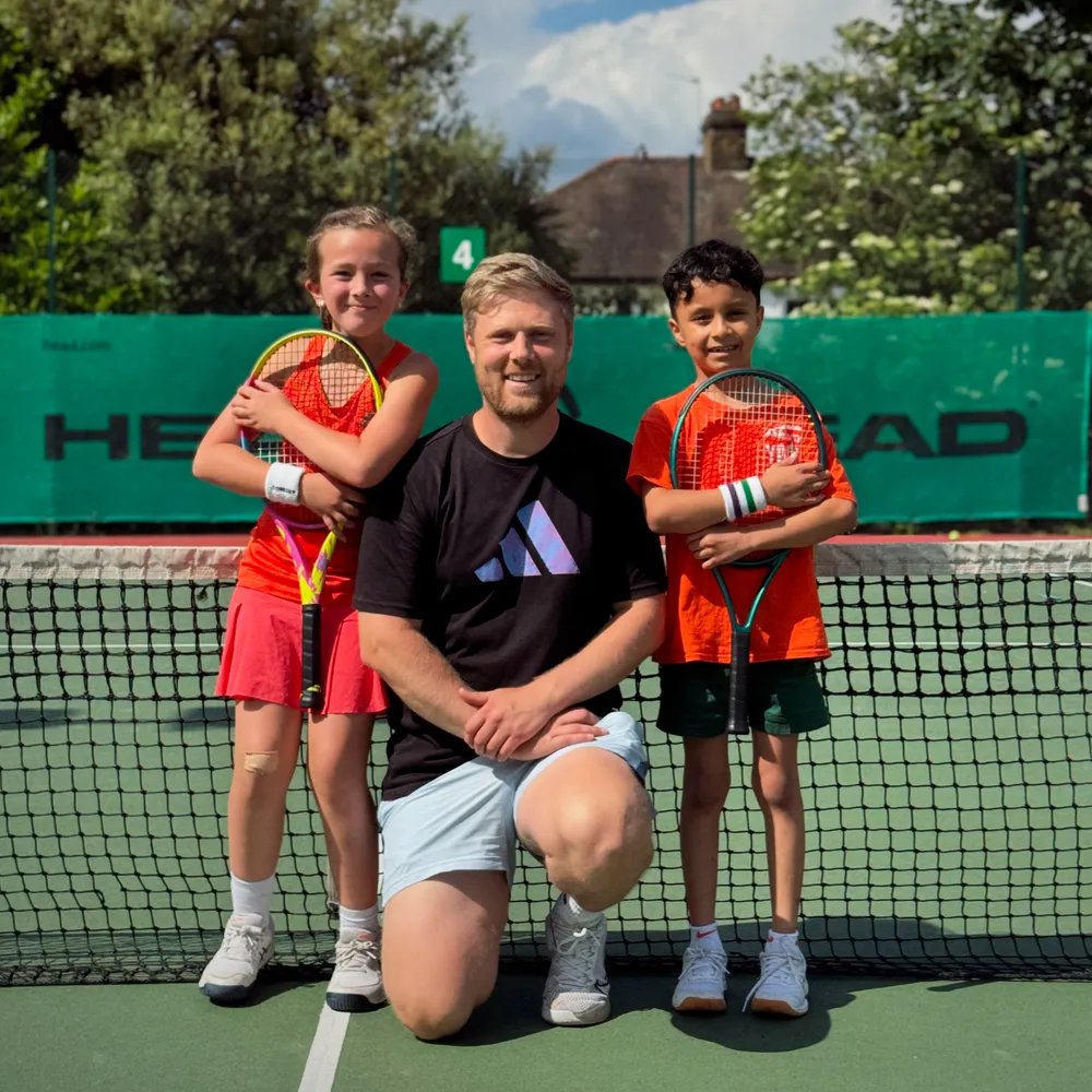James Charman kneeling at the net between two smiling junior players holding tennis rackets