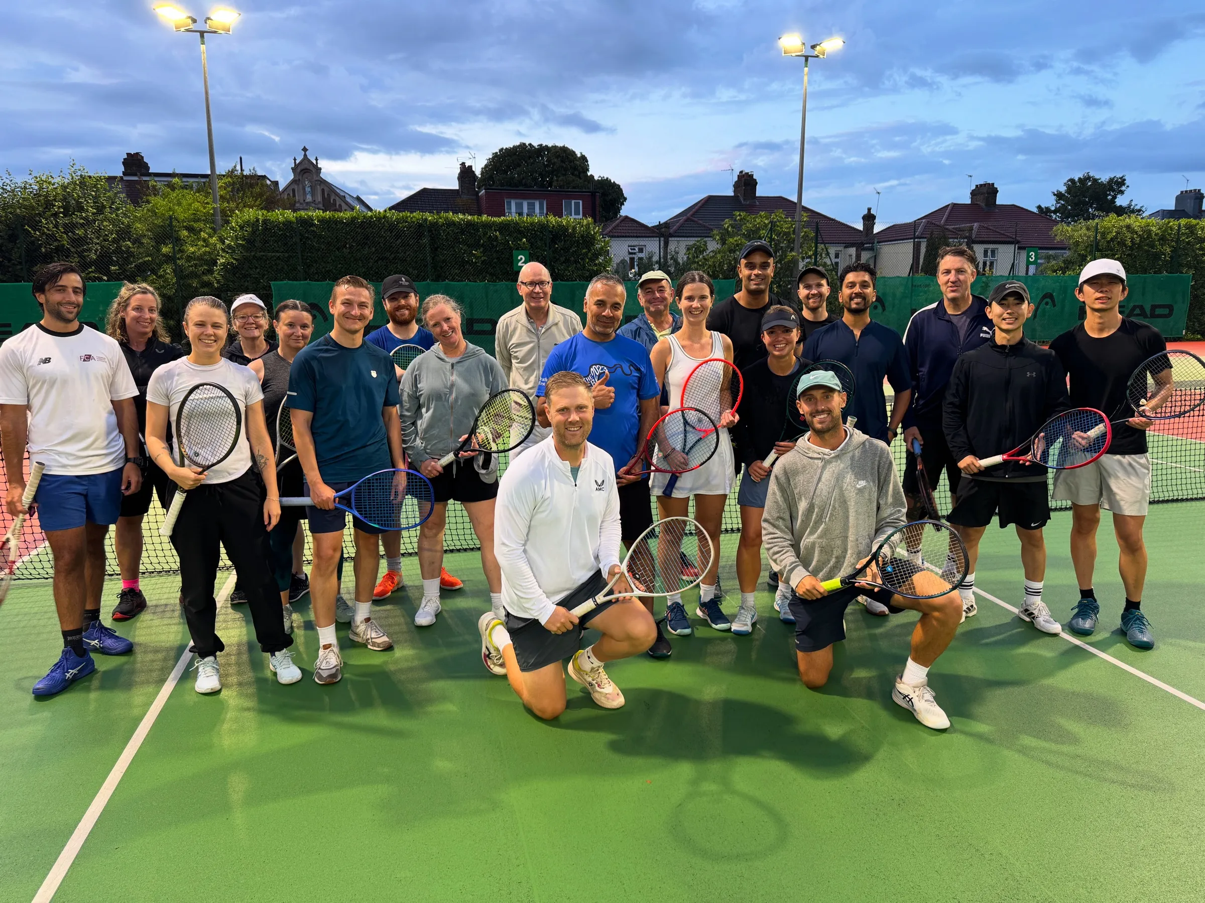 A large group of members gathered on the courts with their rackets under the floodlights at dusk