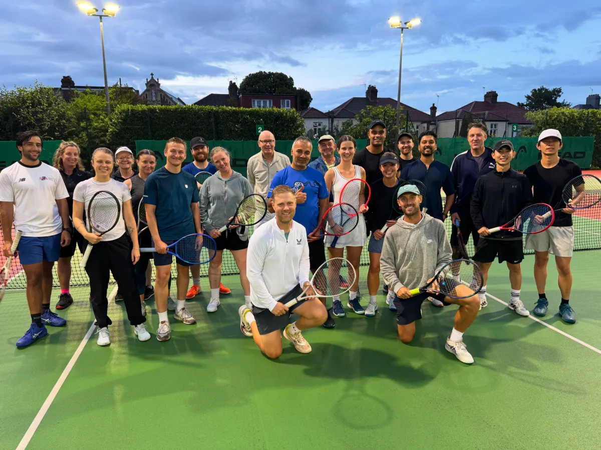 A large group of members with rackets gathered on the courts under the floodlights at dusk