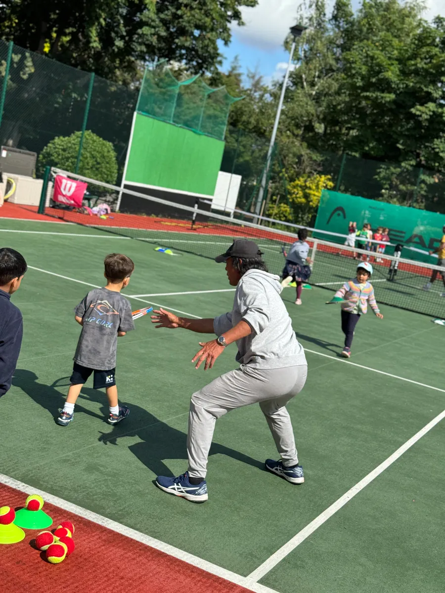 A coach running a junior session on the hard courts, greeting one of the children mid-drill