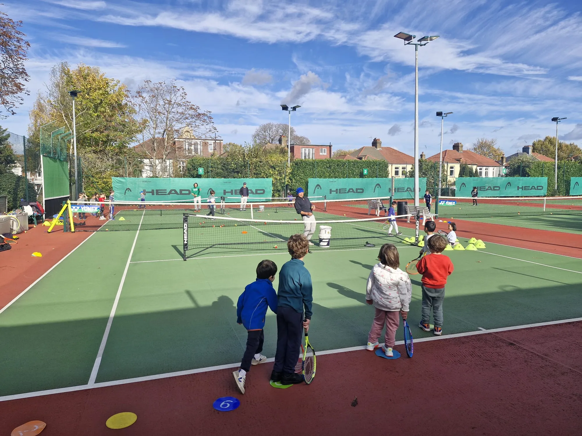 Young children lined up with rackets watching a coach feed balls across a mini net on the club's hard courts